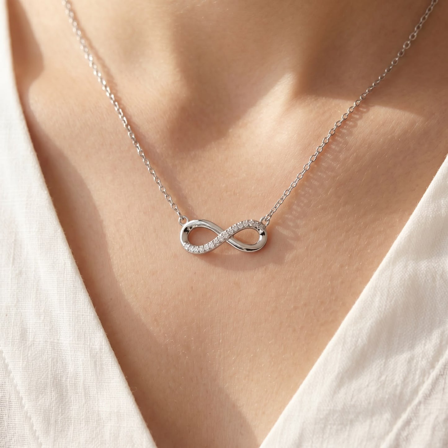 Close-up of a delicate silver necklace featuring a horizontal infinity symbol pendant, half-paved with clear stones and half-polished, worn on a person's neckline.