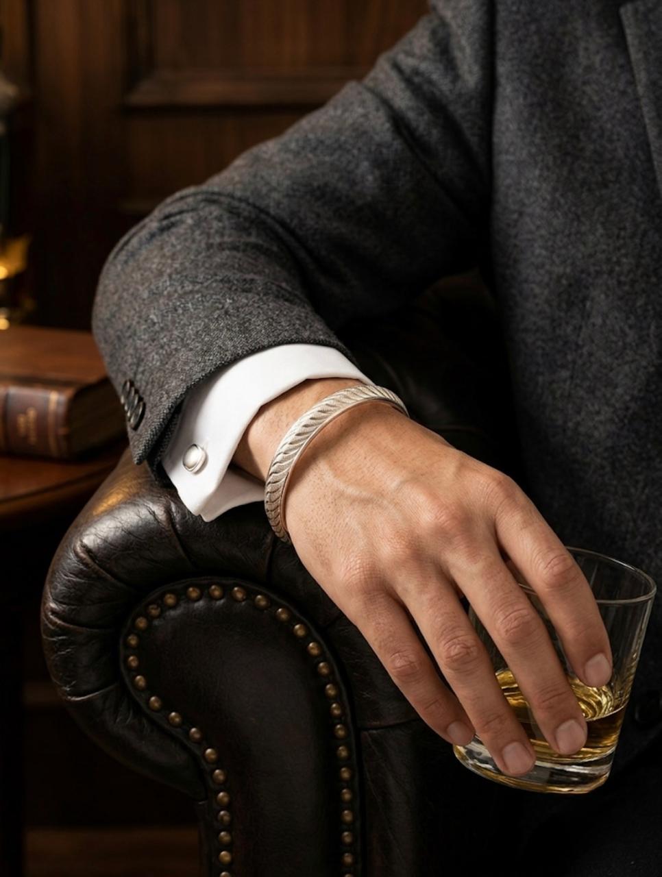 Close-up of a men's textured silver cuff bracelet with carved diagonal grooves, worn on a wrist holding a glass while styled in a charcoal suit.