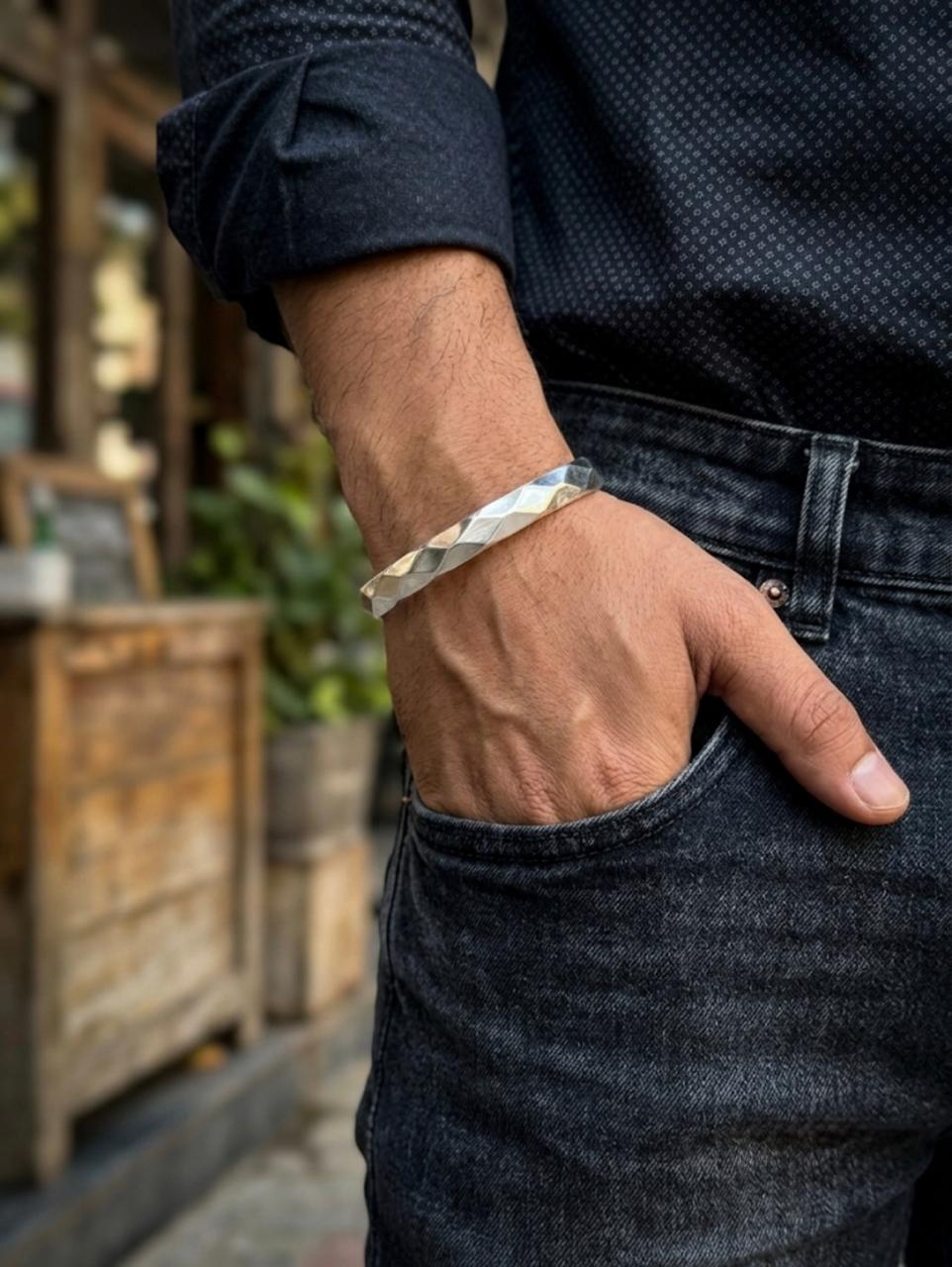 Close-up of a men's silver kada cuff bracelet with a faceted geometric design, worn on a wrist styled with casual dark denim.