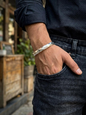 Close-up of a men's silver kada cuff bracelet with a faceted geometric design, worn on a wrist styled with casual dark denim.