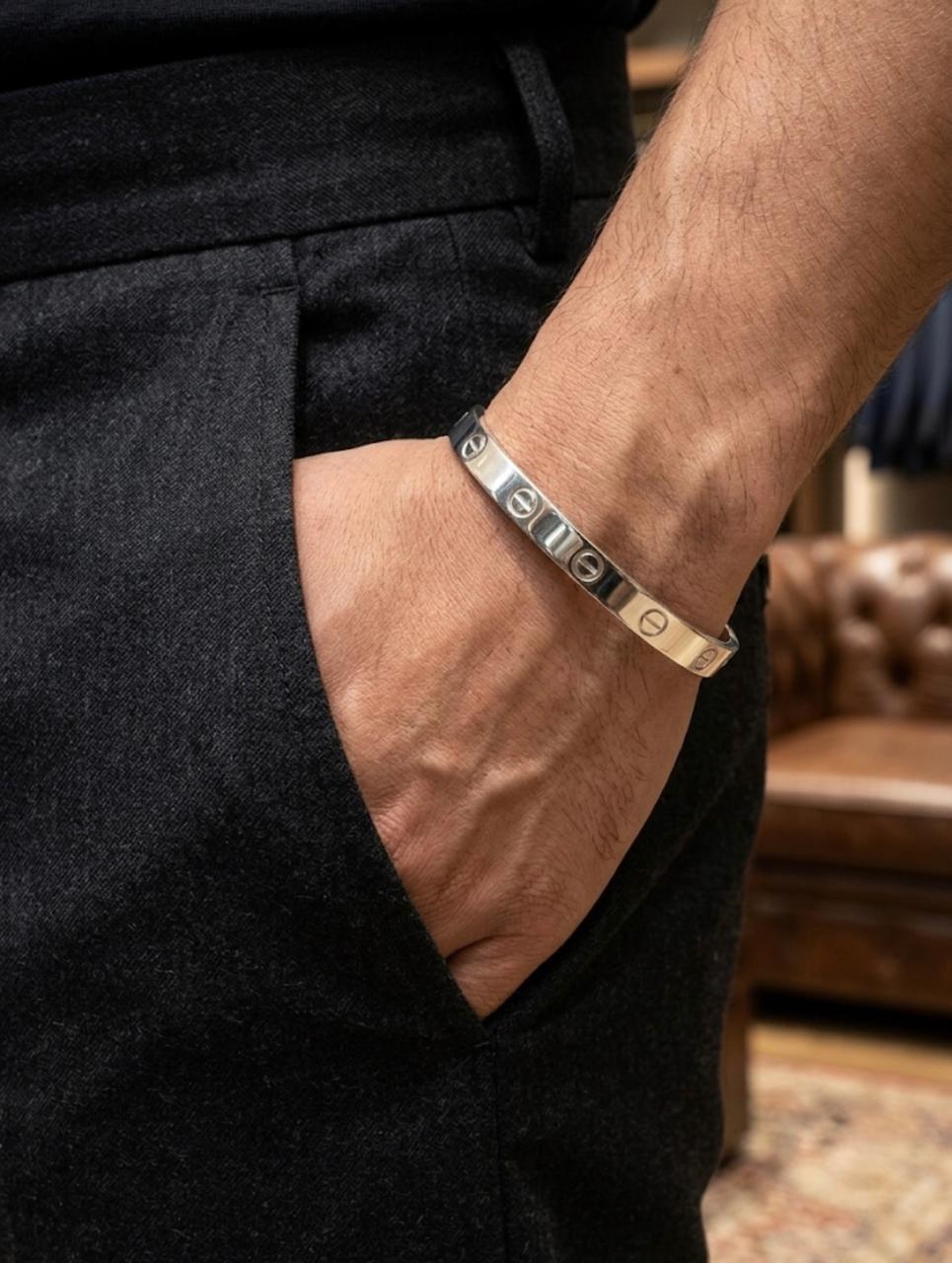 Close-up of a men's polished silver kada cuff bracelet featuring engraved screw motifs, worn on a wrist styled with dark trousers.