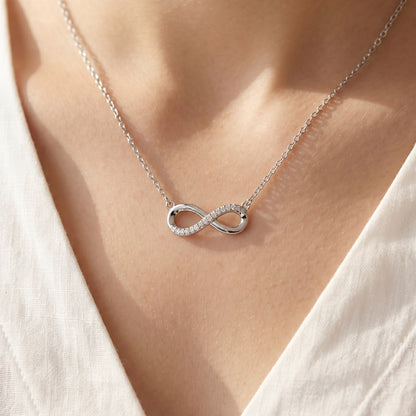 Close-up of a delicate silver necklace featuring a horizontal infinity symbol pendant, half-paved with clear stones and half-polished, worn on a person's neckline.