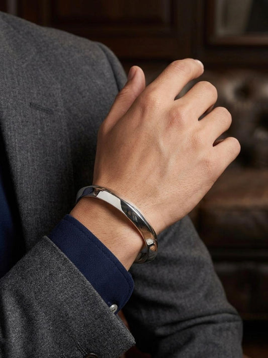 Close-up of a men's solid polished silver cuff bracelet worn on the wrist, styled with a formal grey blazer and blue shirt.