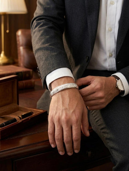 Close-up of a men's silver cuff bracelet featuring an engraved geometric chevron pattern, worn on the wrist in a formal suit setting.