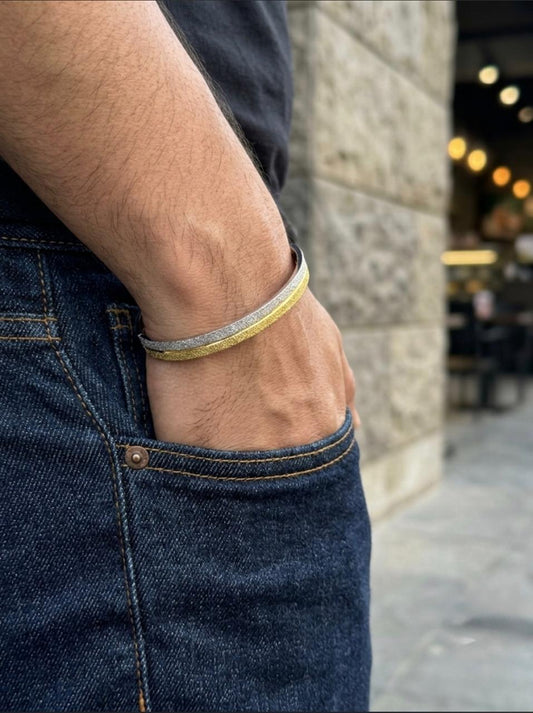 Close-up of a men's dual-tone silver kada cuff bracelet with a frosted silver and gold-tone textured finish, worn on a wrist styled with dark denim.