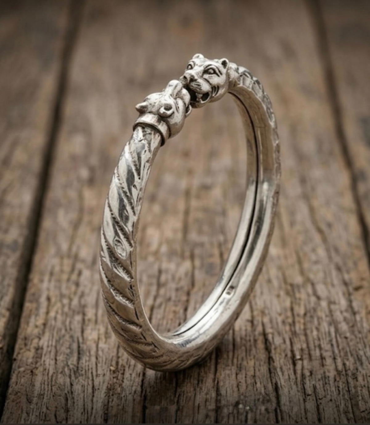 925 silver men's kada with sculpted twin lion head motifs and textured engraved detailing, displayed upright on a wooden surface.